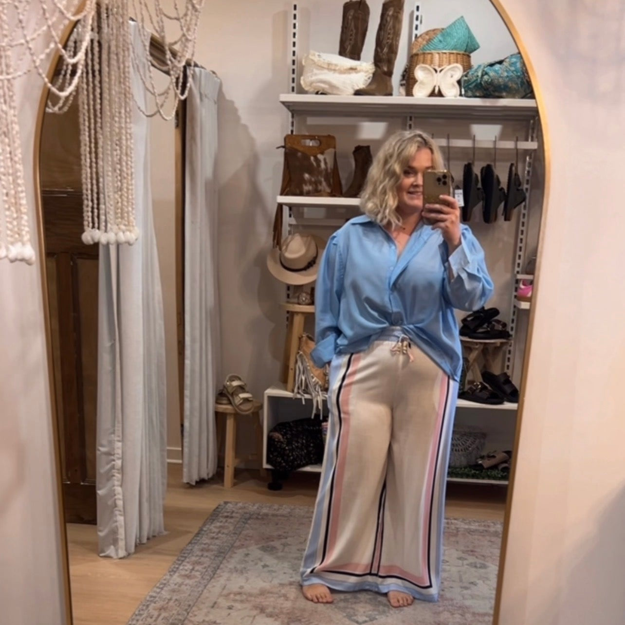 Person taking a mirror selfie wearing a blue shirt and beige pants in a room with a rug and shelves.
